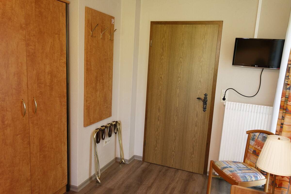 Une pièce comprenant une armoire en bois, une porte avec un porte-manteau, un téléviseur fixé au mur et une chaise avec une lampe.