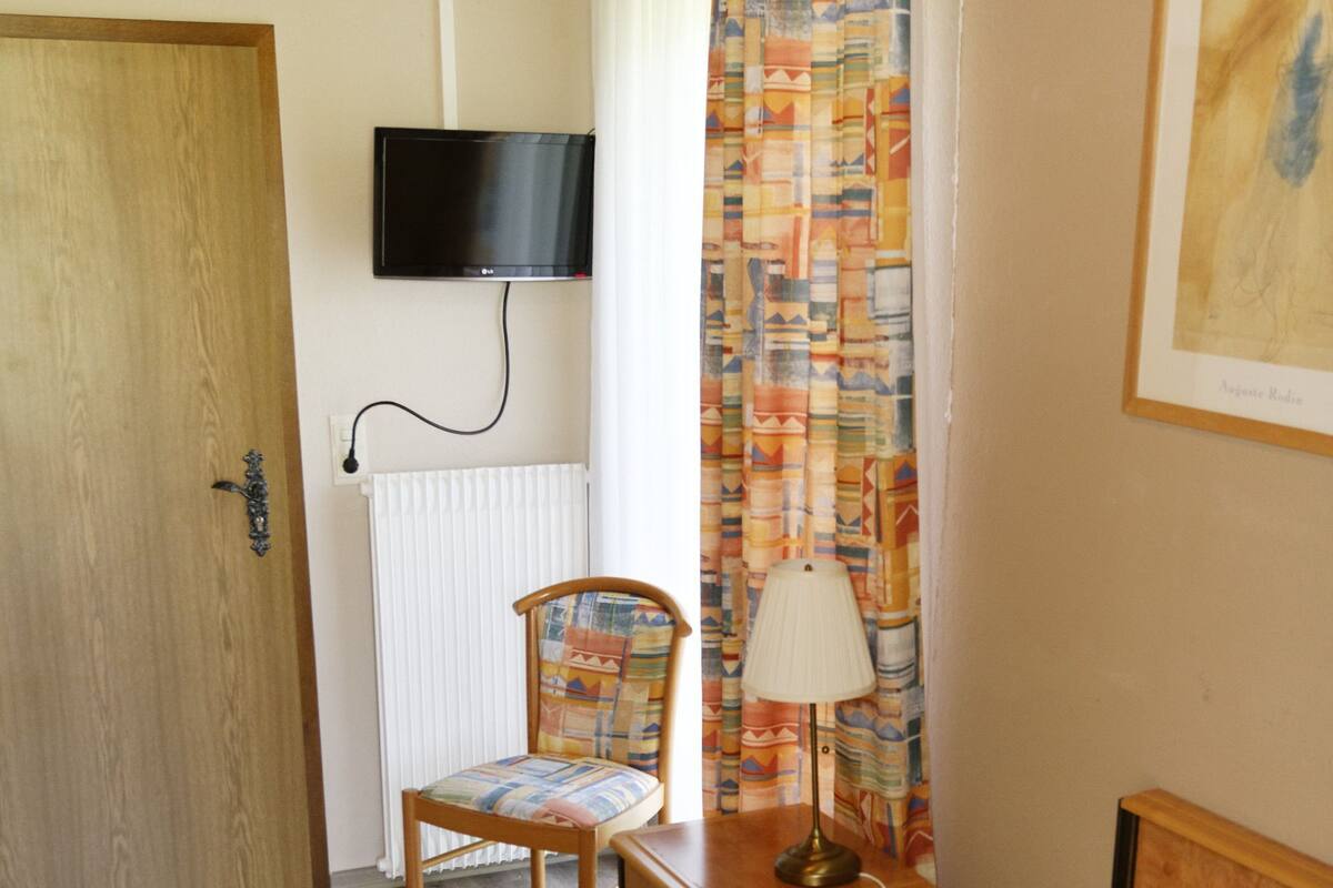 Une chambre à coucher comprenant un lit, une chaise, une table avec une lampe, un téléviseur fixé au mur et une porte.