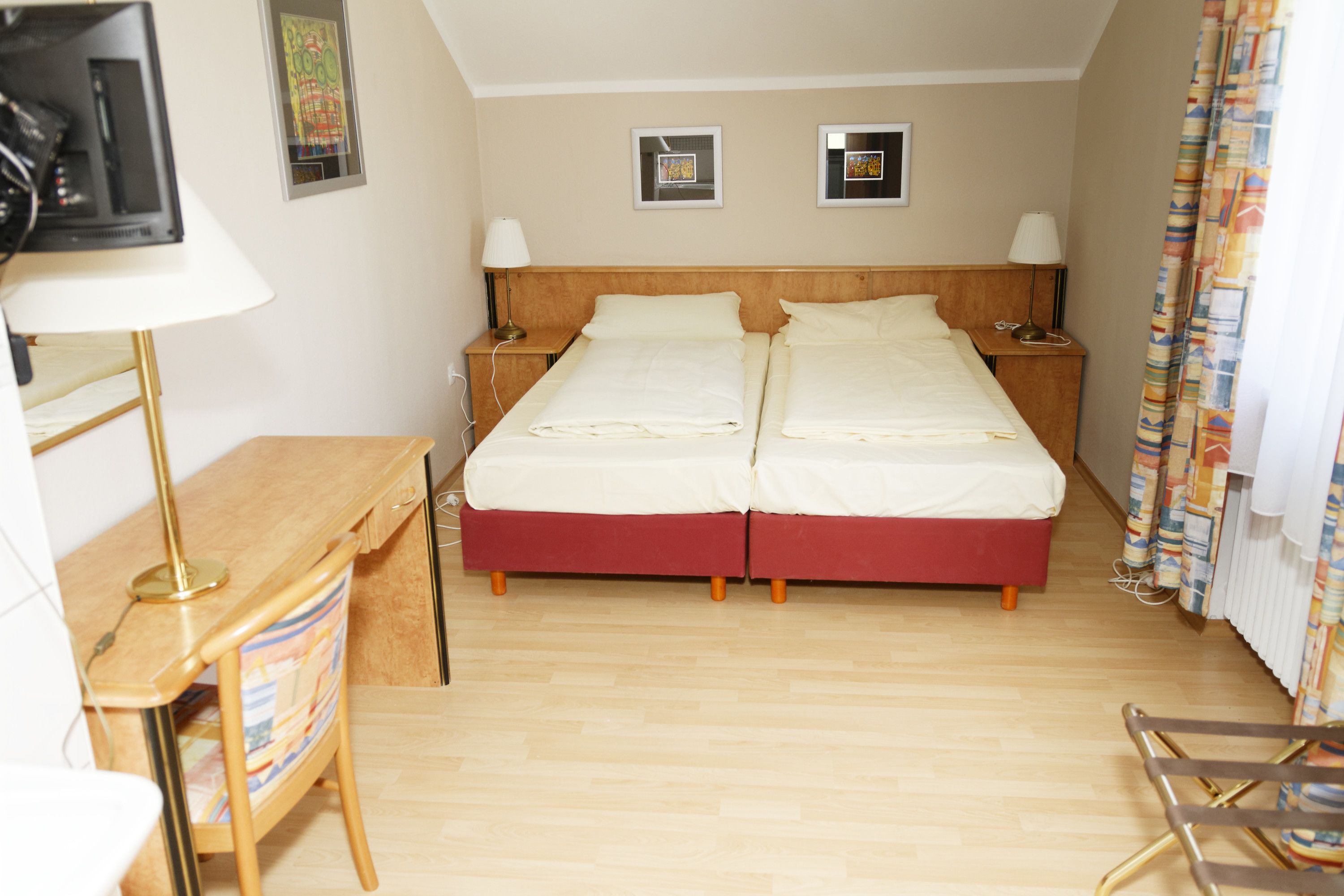 Ein Hotelzimmer mit einem Doppelbett, einem Schreibtisch mit Stuhl, einem Fernseher, zwei Nachttischen mit Lampen und einem Fenster mit Vorhängen.