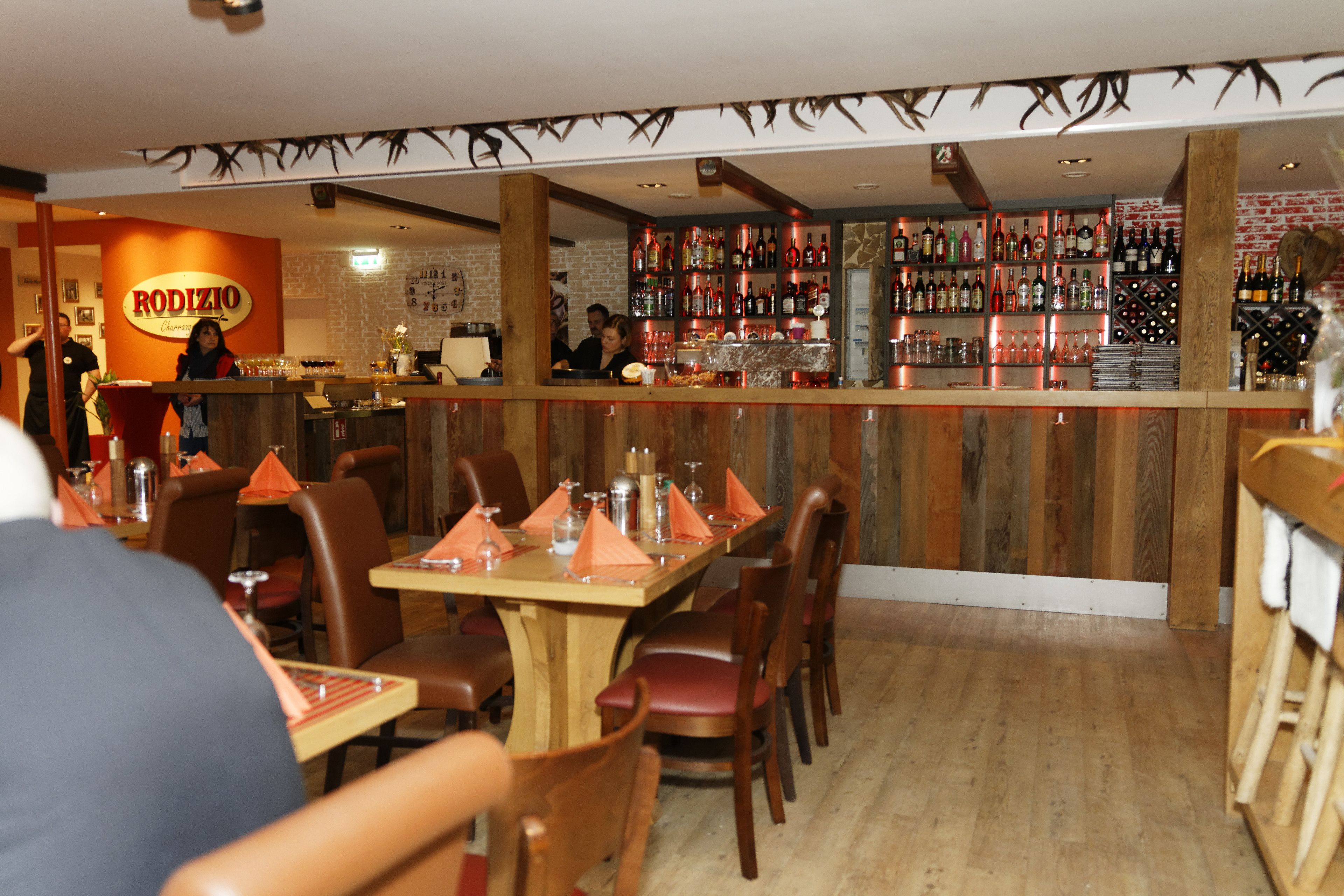 A restaurant interior with wooden tables and chairs, a bar area with shelves of bottles, and a sign for 'RODIZIO'.