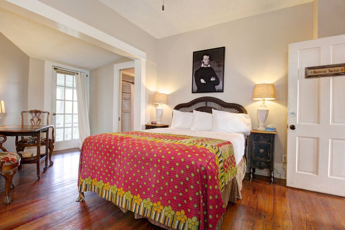 A bedroom with a large bed, two bedside tables with lamps, a chair, a door labeled "Bowie Suite," and a framed picture on the wall.