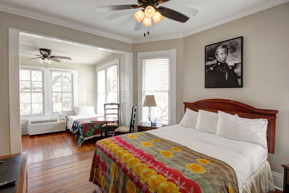 A bedroom with a large bed, a ceiling fan, two windows with blinds, a chair, and a bedside table with a lamp.