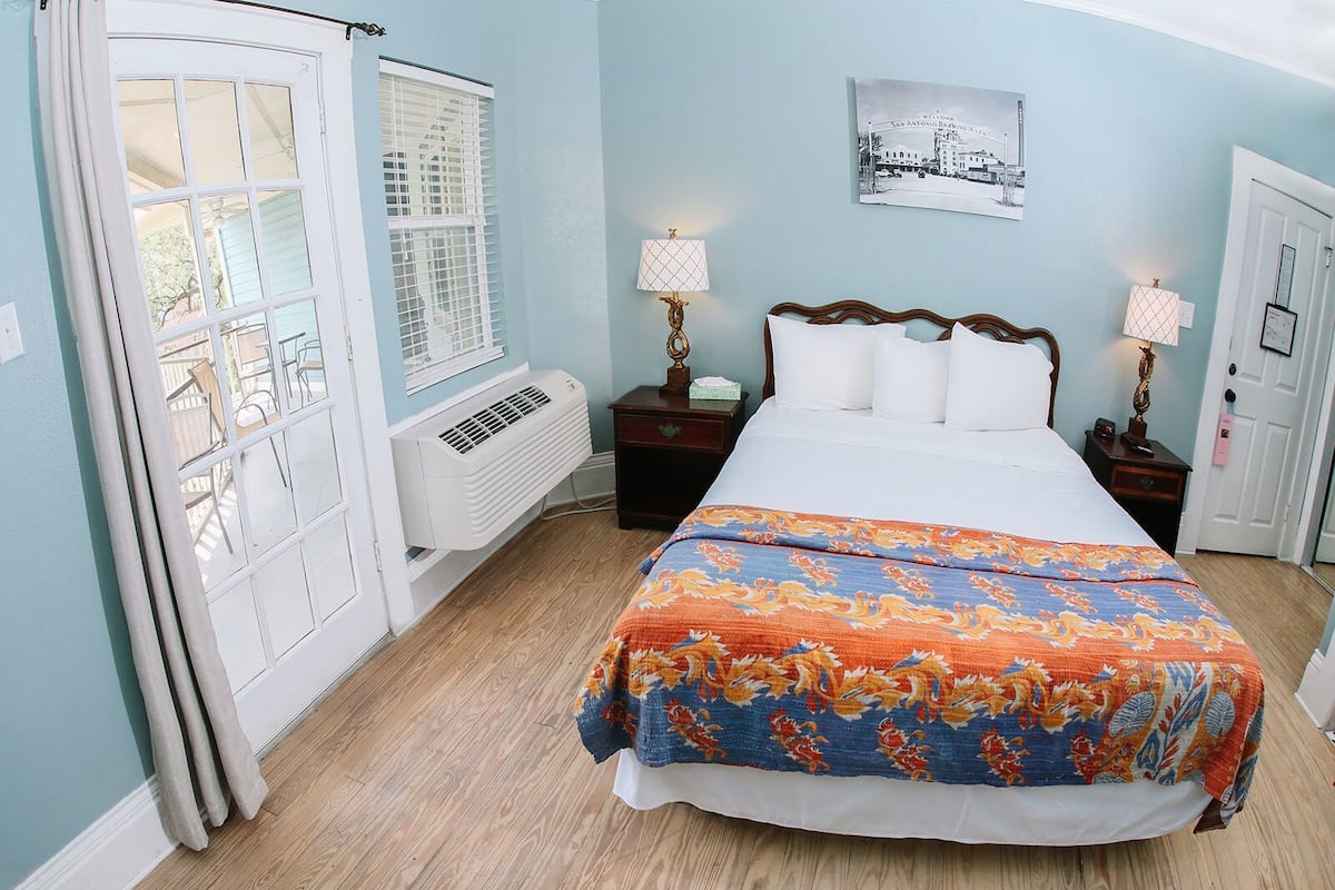 A bedroom with a bed, two bedside tables, a wall-mounted lamp, a radiator, and a window with blinds.
