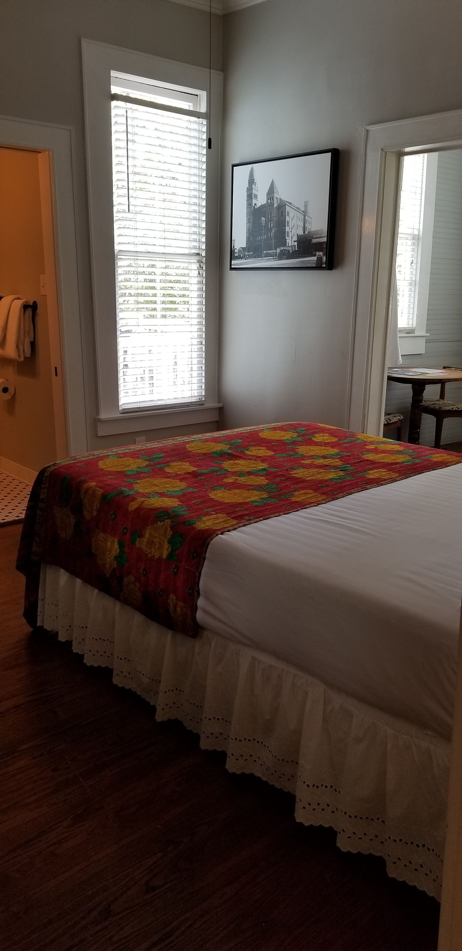 A bedroom with a bed, a window with blinds, a framed picture on the wall, and a door.