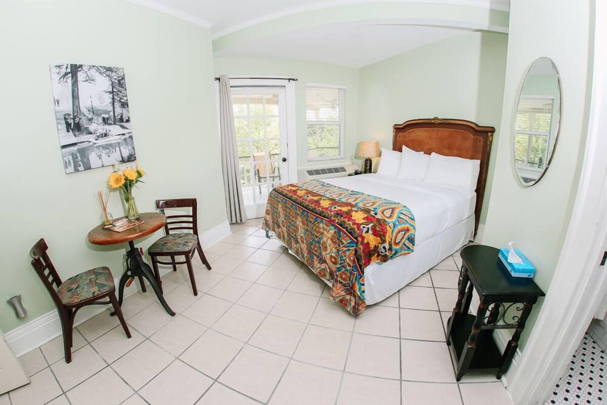 A bedroom with a bed, a chair, a small table with a vase of flowers, and a window.