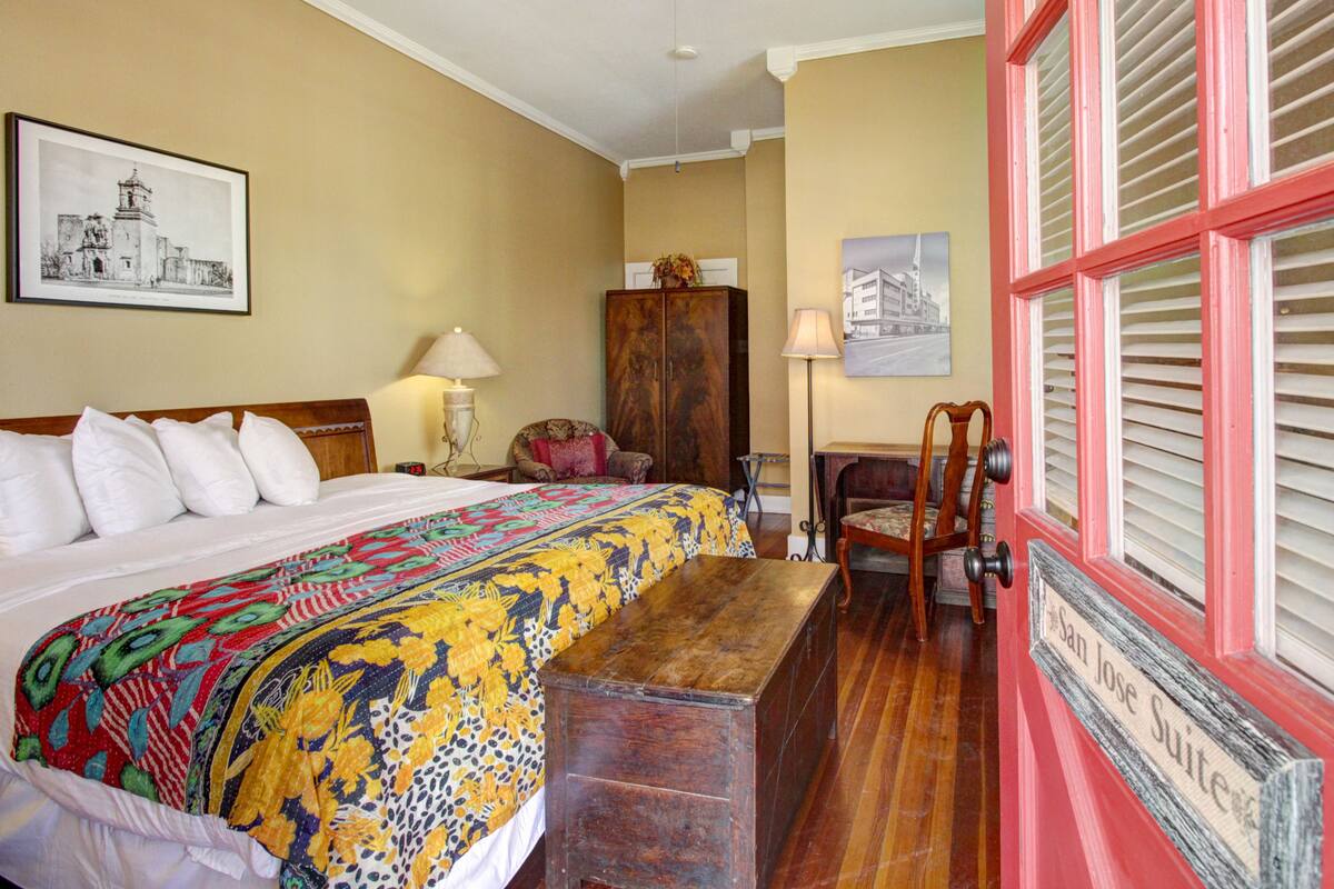 A hotel room with a large bed, a wooden bench, a desk, and a chair. There is a cabinet and a lamp on the desk.