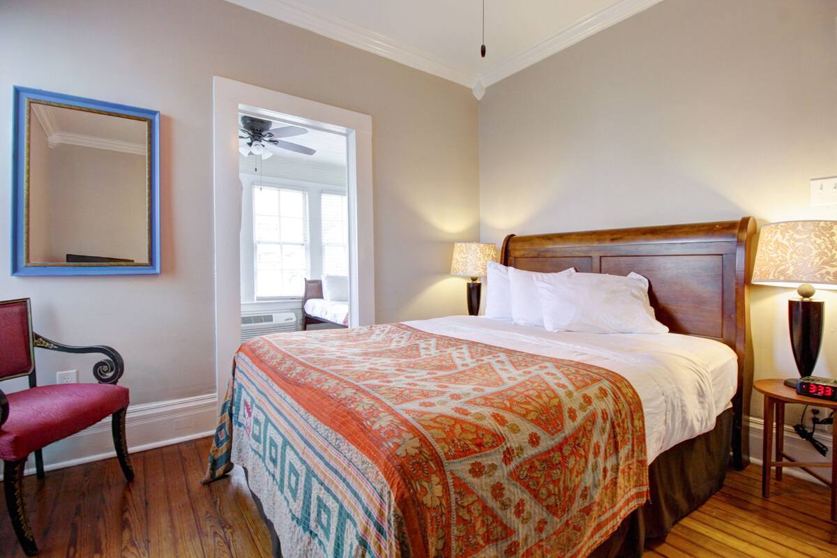 A neatly made bed with a patterned bedspread, a wooden headboard, and two bedside lamps. There is a chair with a red cushion and a large mirror on the wall.