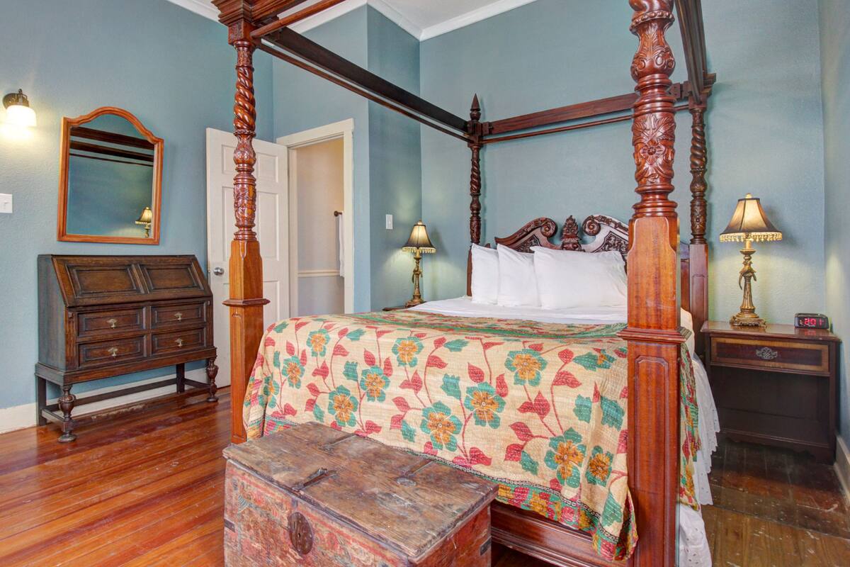 A four-poster bed with a floral bedspread, a wooden nightstand with a lamp, a dresser, and a mirror on the wall.
