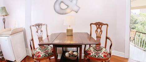 Un área de comedor con una mesa de madera y dos sillas con tapicería floral, una letra decorativa colgada en la pared y una lámpara al lado.
