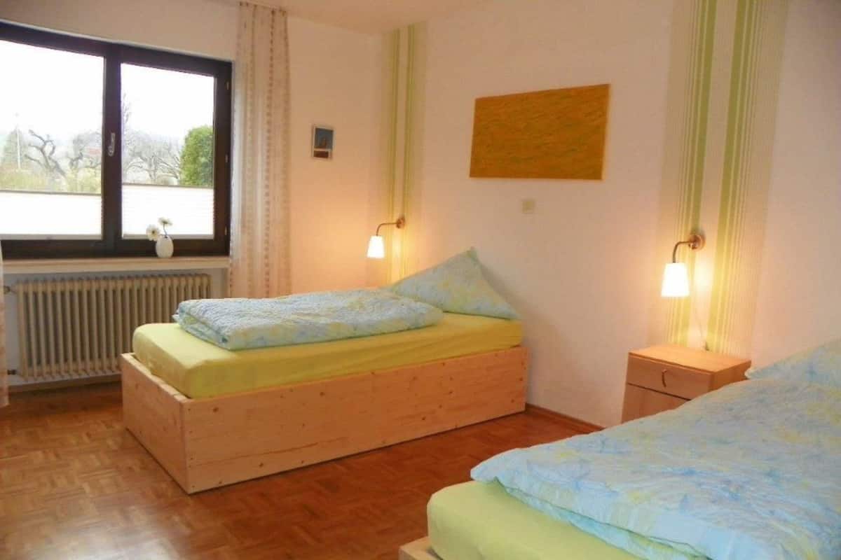 A room with two wooden beds, a window with curtains, a radiator, and wall-mounted lamps.
