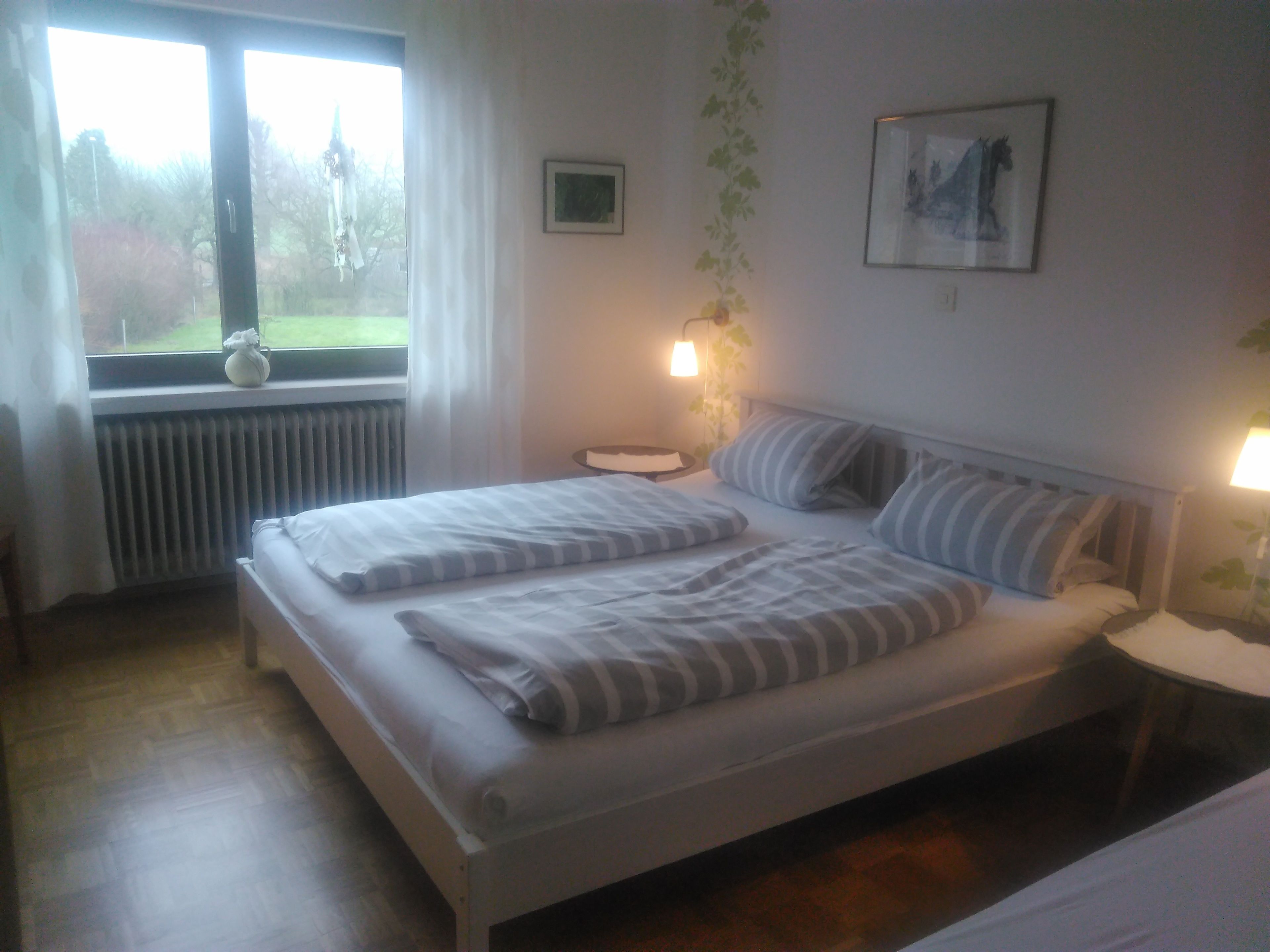 Ein ordentlich bezogenes Bett mit Streifenmuster, ein Nachttisch mit Lampe, ein Fenster mit Vorhängen und ein gerahmtes Bild an der Wand.