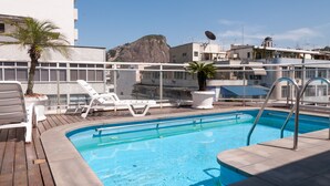 Una piscina en la azotea con sillas de descanso blancas, una palmera y vistas a una colina rocosa.