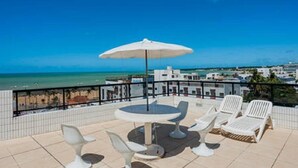 Una terraza con una mesa redonda y sillas blancas, con vistas a la playa y al paisaje urbano.