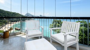 Varanda com duas cadeiras brancas e uma mesa pequena, com vista para uma praia e oceano.