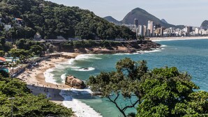 A coastal cityscape with a beach, rocky shoreline, and a bridge connecting to a hillside. - Omar do Rio - Apartamento V210 (Rio de Janeiro)