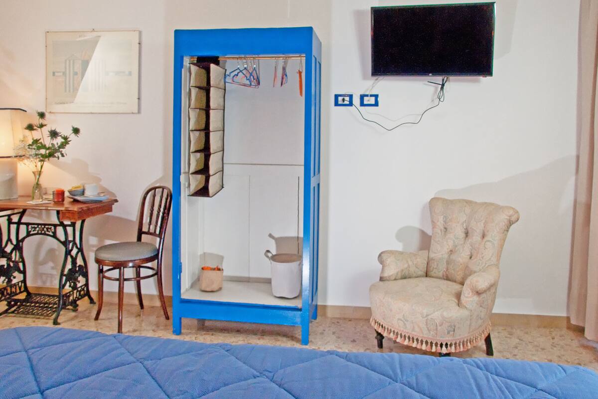 A room with a blue wardrobe, a beige armchair, a wooden table with a chair, a lamp, and a framed picture on the wall.