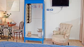 Una stanza con un armadio blu, una poltrona beige, un tavolo di legno con una sedia, una lampada e un quadro appeso al muro.