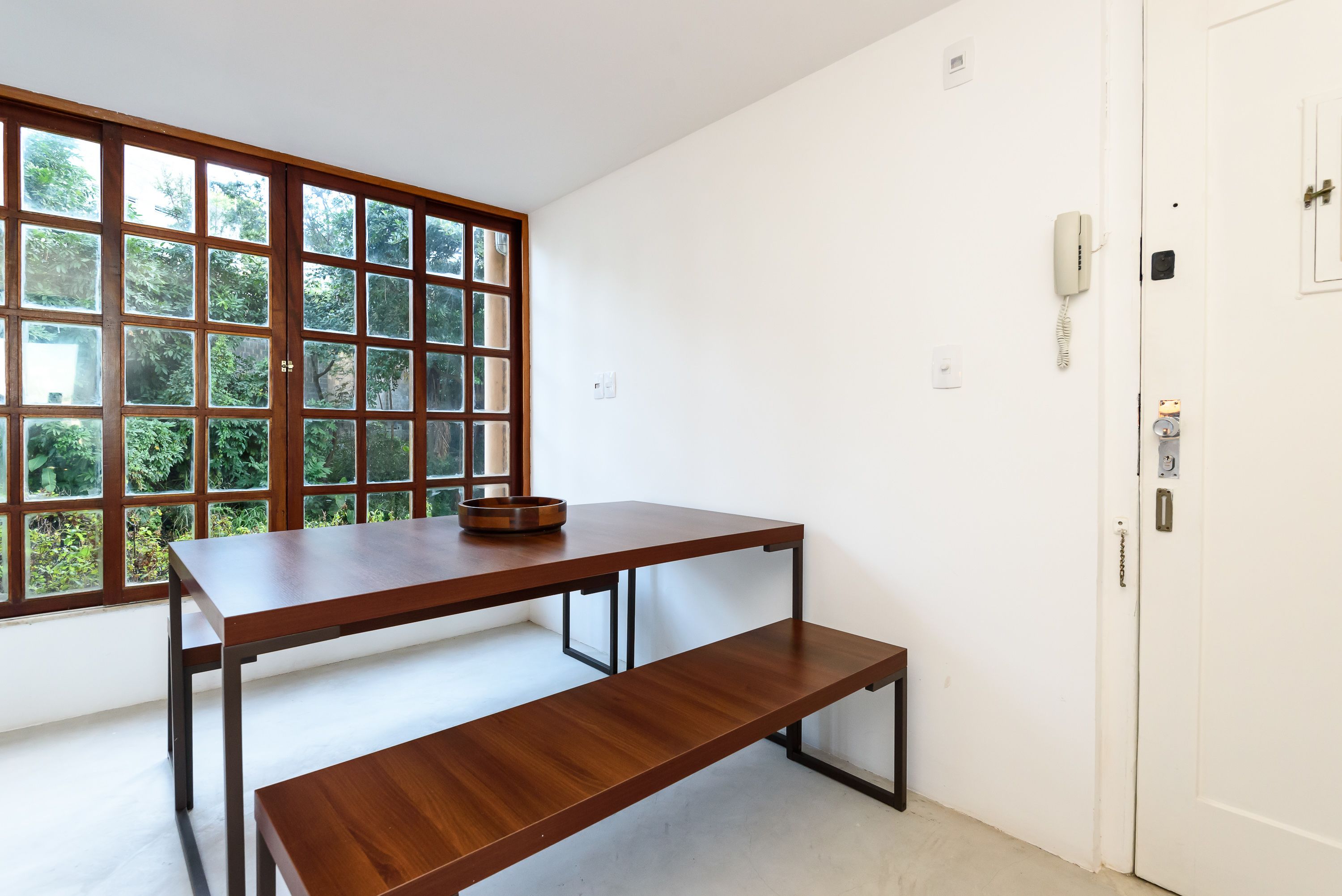Una habitación con una mesa de madera y un banco, una ventana con vistas a zonas verdes, y una puerta blanca con un teléfono.