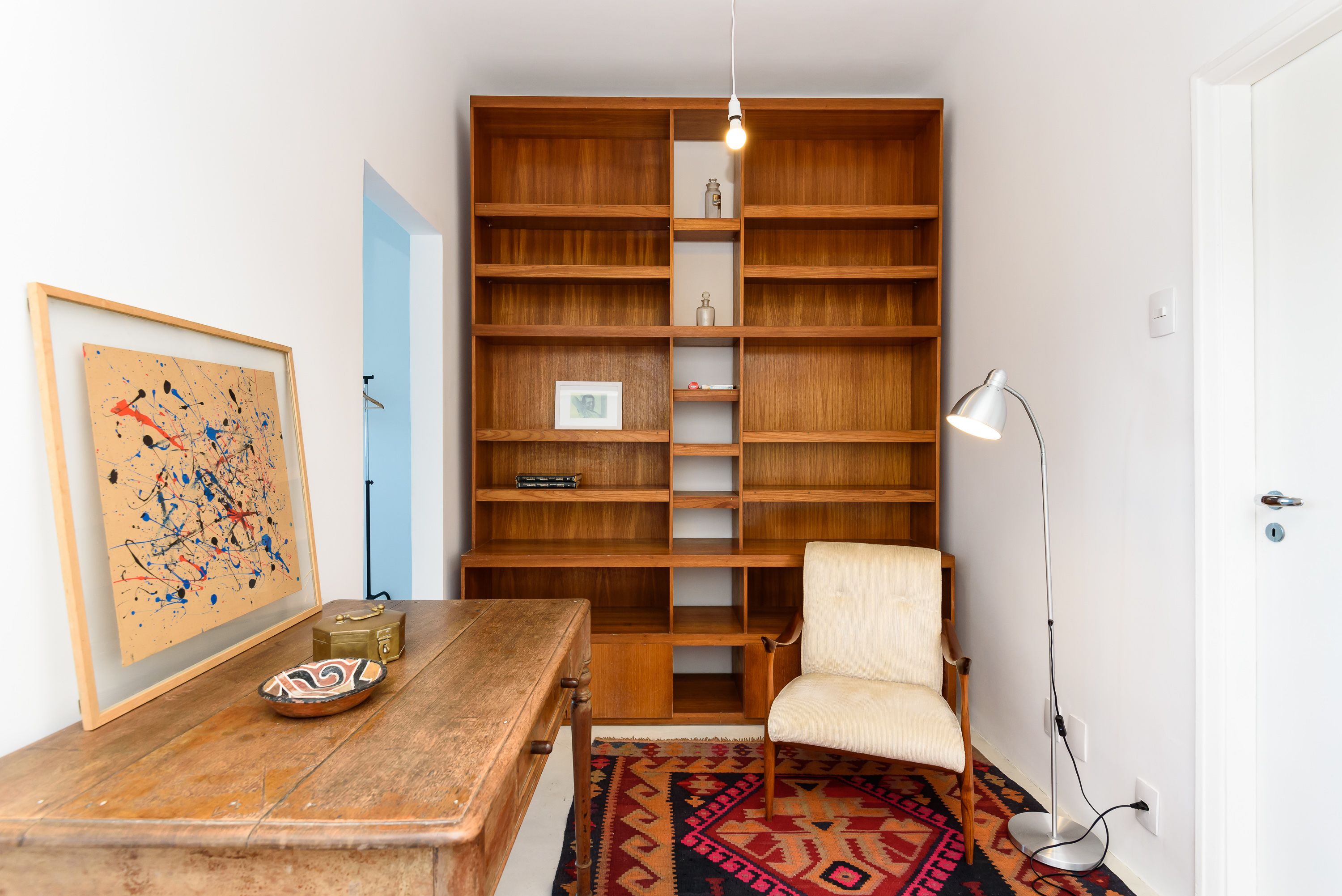 A room with a wooden bookshelf, a white chair, a wooden table, and a floor lamp.