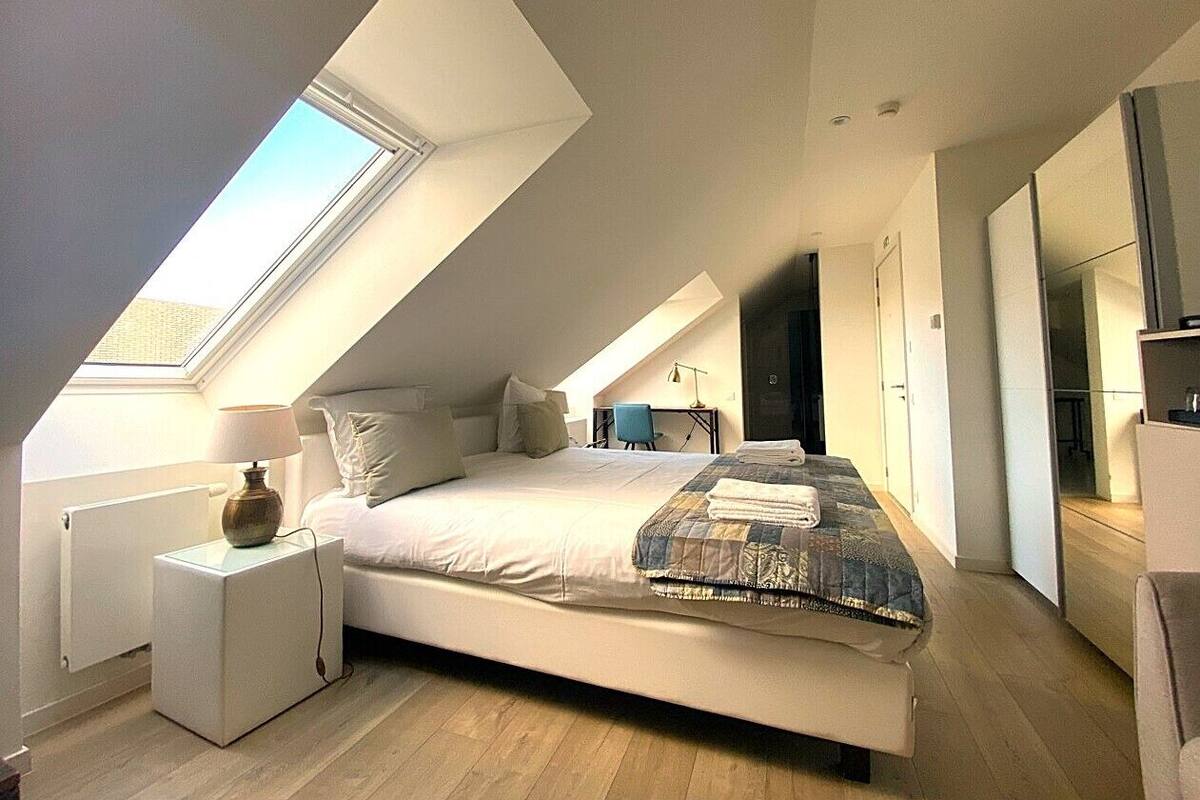 Quarto moderno com claraboia, cama com colcha, mesa de cabeceira e uma cadeira.
