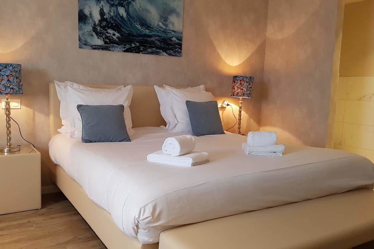 Cama bem arrumada com lençóis brancos, dois travesseiros e duas toalhas enroladas. Há duas luminárias de cabeceira com motivos florais nas mesas de apoio.