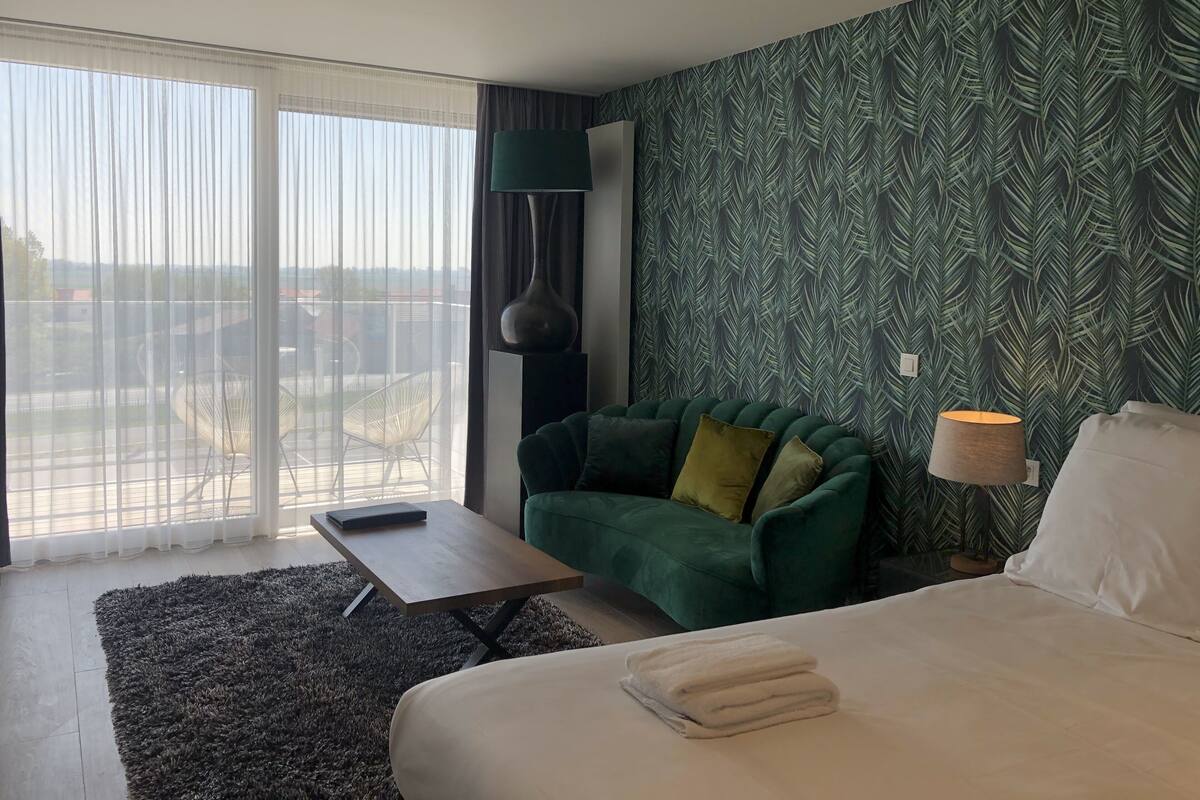 Quarto de hotel moderno com uma cama grande, uma poltrona verde, uma mesa pequena e vista da cidade através de grandes janelas.