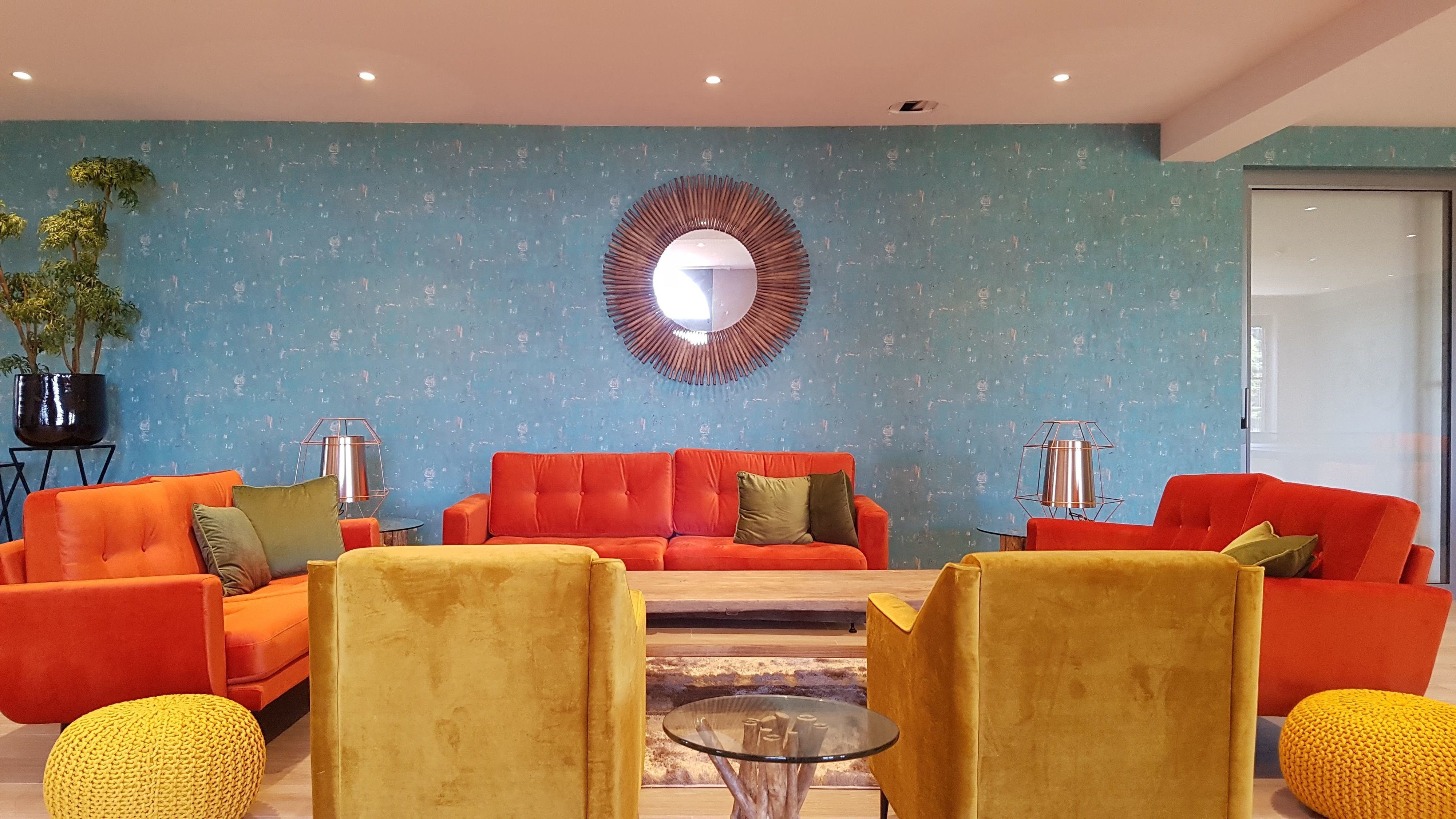 Een moderne woonkamer met een blauwe, gestructureerde muur, een grote ronde spiegel en twee rode banken met gele en groene kussens.