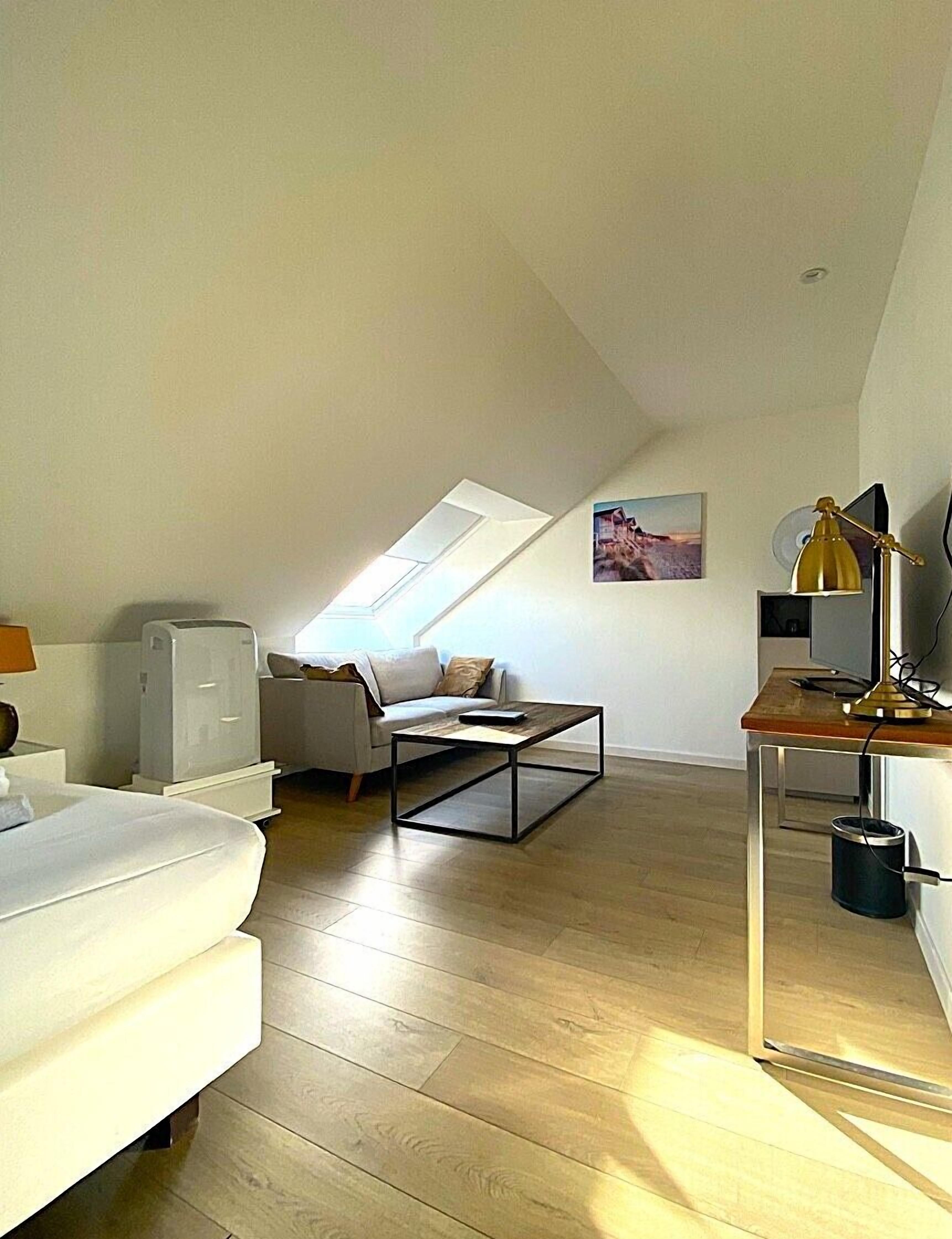 A modern loft apartment with a sloped ceiling, a white sofa, a coffee table, a desk with a lamp, and a painting on the wall.