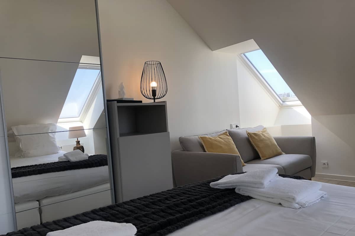 Quarto de hotel moderno com clarabóia, cama com lençóis brancos, sofá cinza com almofadas, mesa lateral e um espelho.