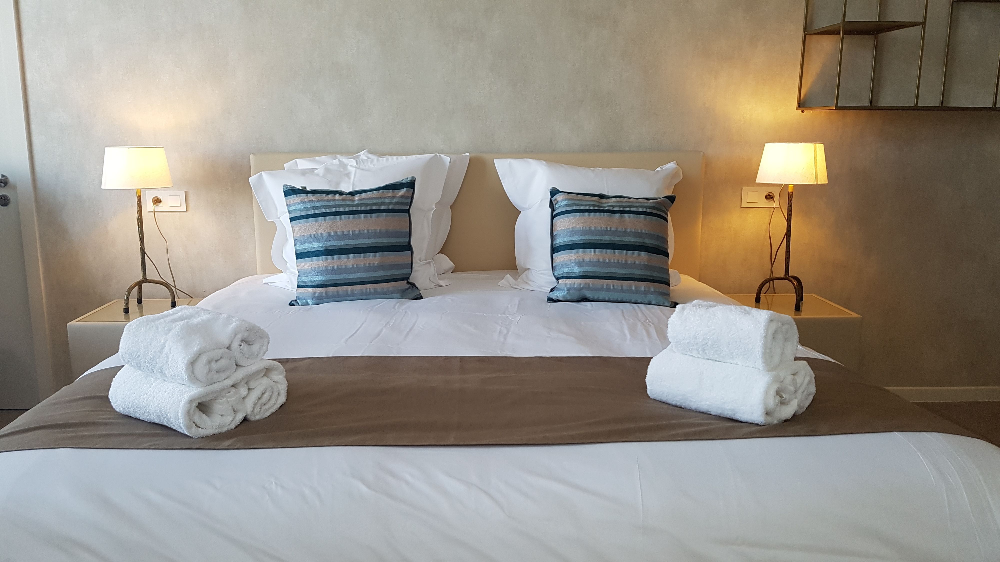 A neatly made bed with white linens, two striped pillows, and two rolled towels on the side.
