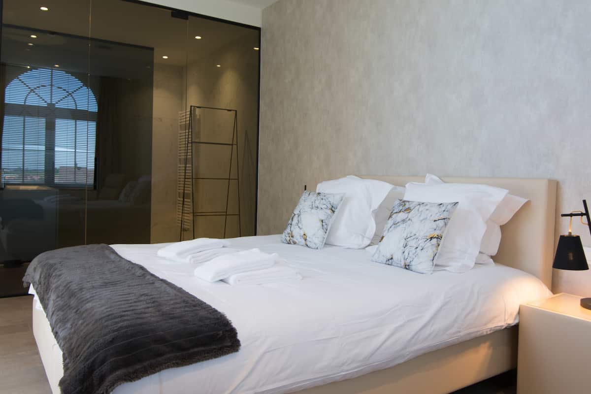 Quarto de hotel moderno com uma cama grande, chuveiro com box de vidro e vista para o horizonte urbano.