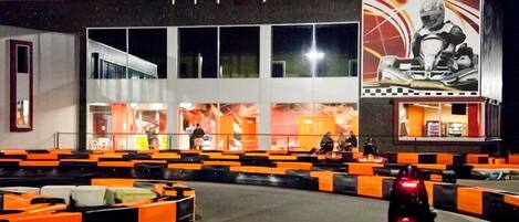 Una pista de karts con barreras naranja y negro, un kart con el número 4, y un edificio con grandes ventanales y un mural de un piloto de karts.