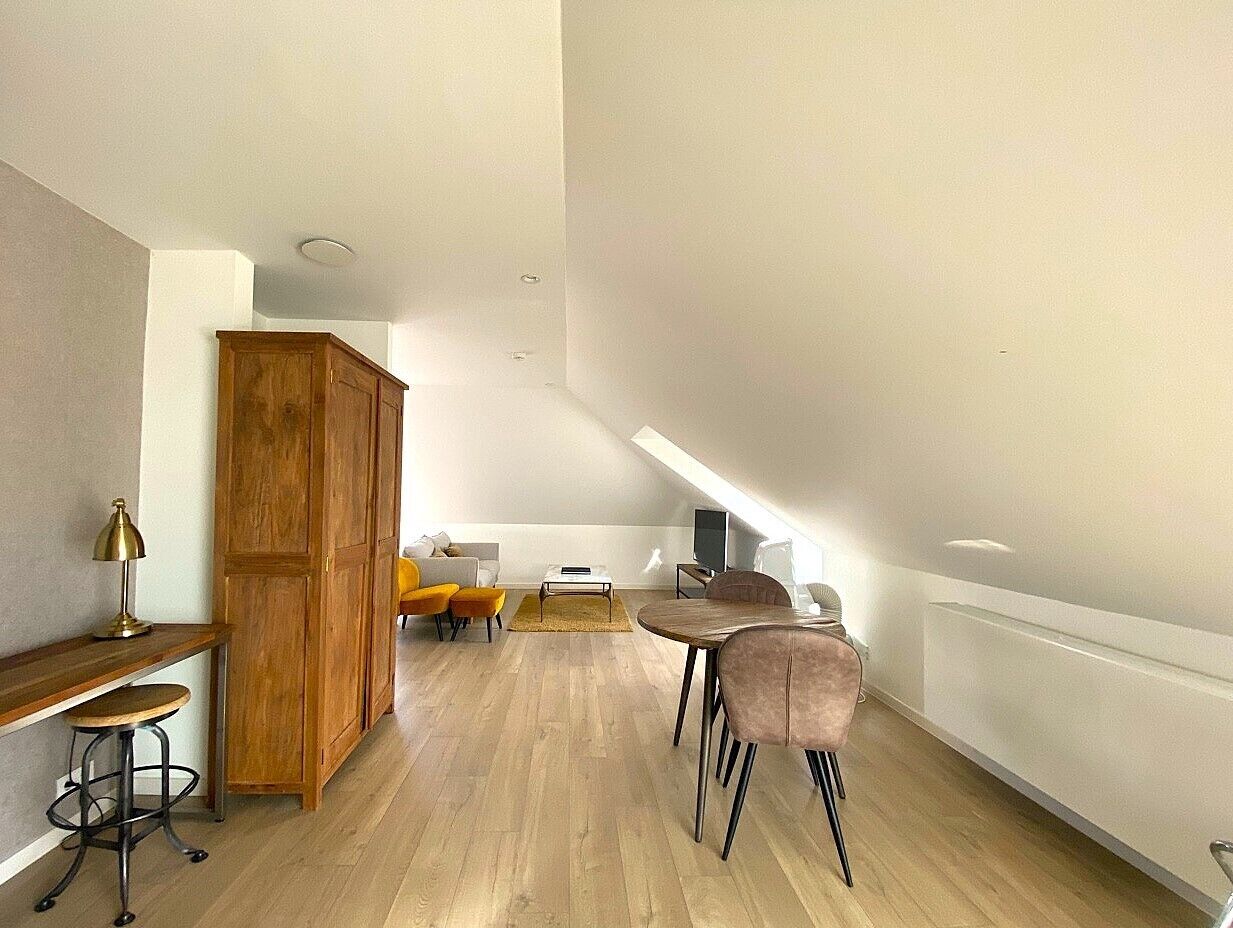 A spacious loft with a high ceiling, wooden flooring, a dining area with a table and chairs, a sofa, and a wooden cabinet.