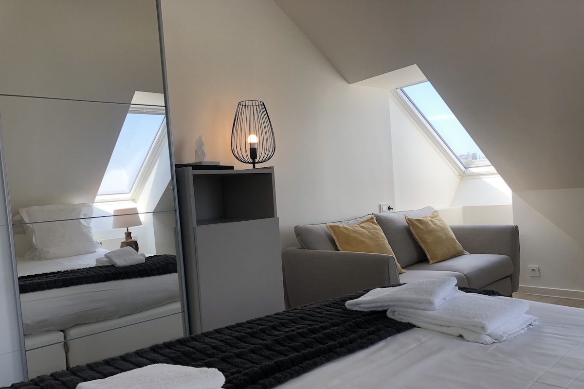 A modern hotel room with a skylight, a bed with white linens, a grey sofa with cushions, a side table, and a mirror.