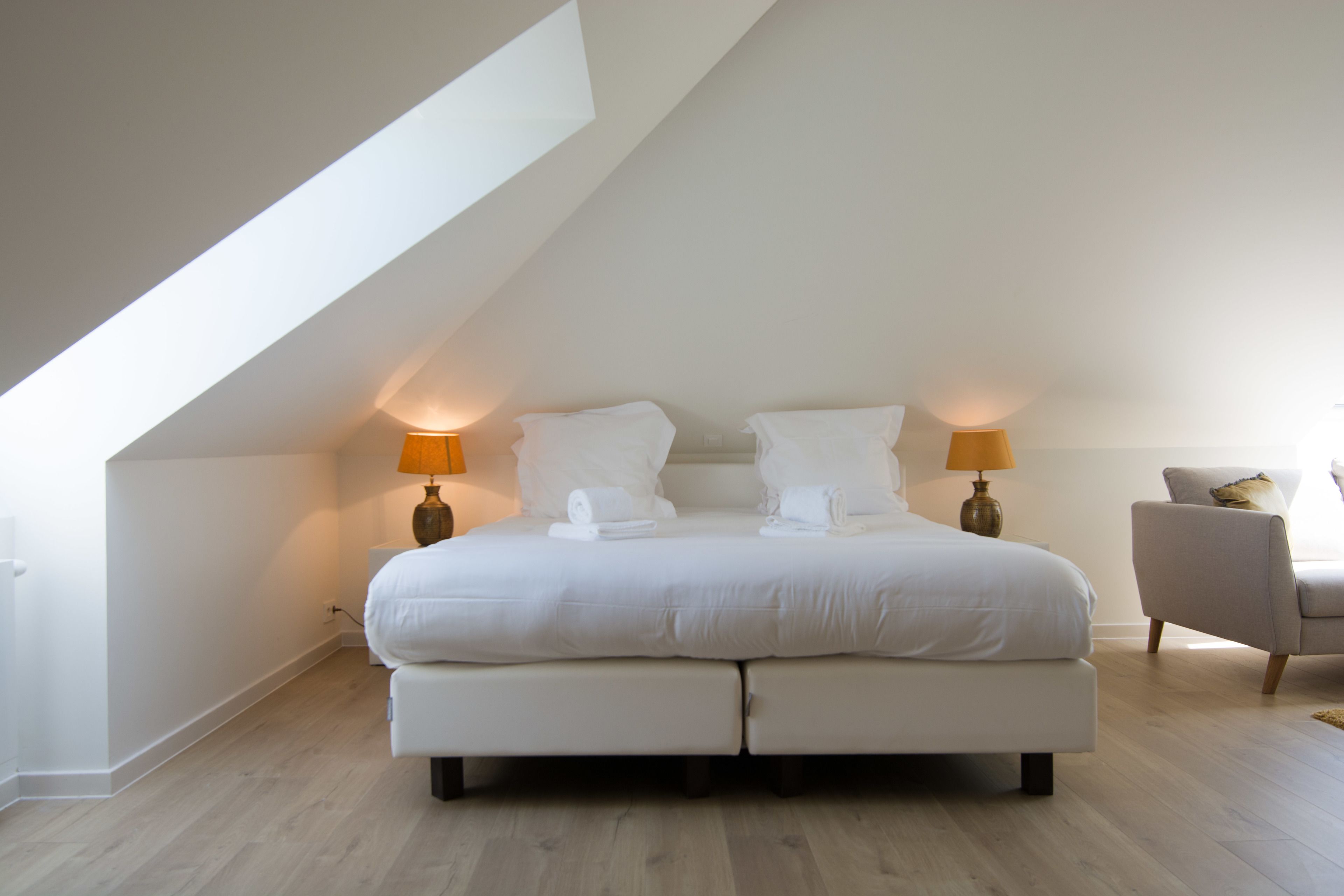 A modern bedroom with a sloped ceiling, a large bed with white linens, two bedside lamps, and a chair.