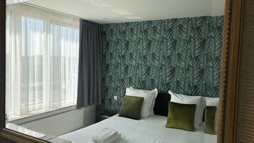 Habitación de hotel con una cama grande, una ventana con cortinas translúcidas, un papel tapiz estampado y un espejo con marco de madera.