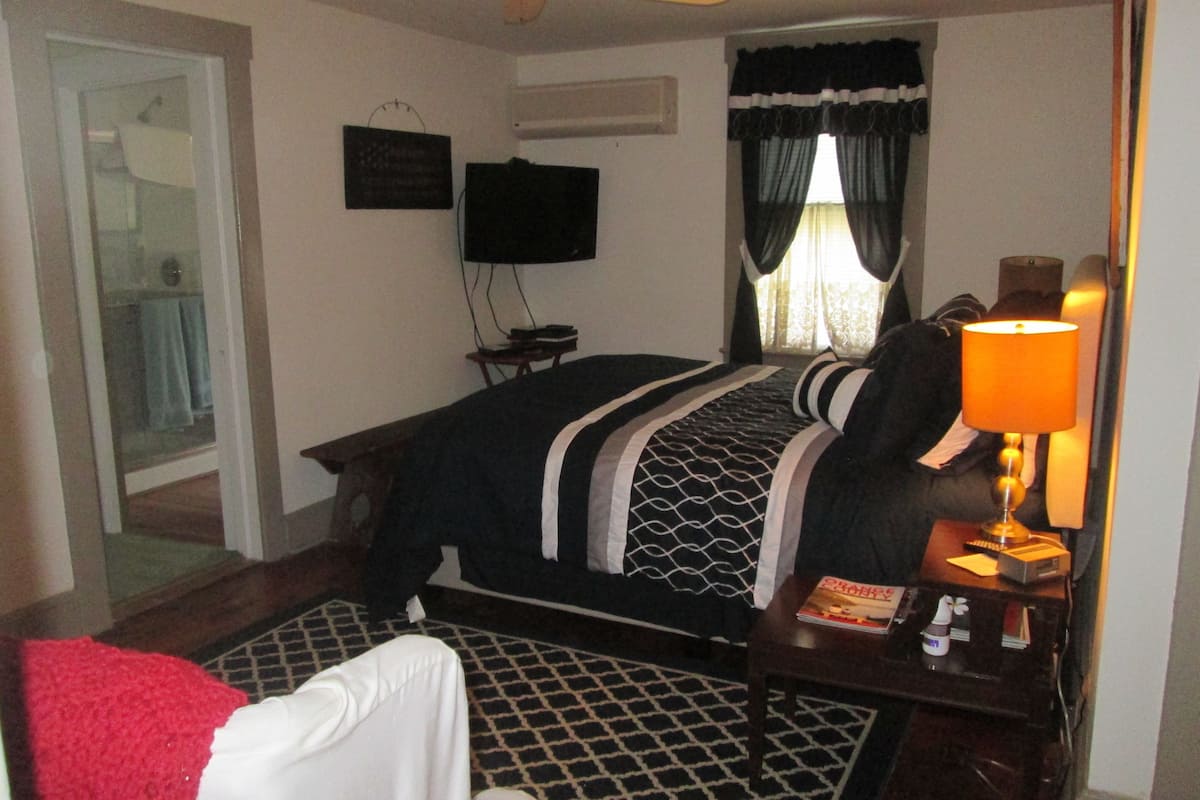 A hotel room with a bed, a desk with a lamp, a TV on the wall, and a window with curtains.