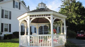 A white gazebo with a roof, surrounded by a white fence, and located in a grassy area with a house in the background. - Buck's Homestead B&B (Montgomery)