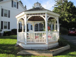 A white gazebo with a roof, surrounded by a white fence, and located in a grassy area with a house in the background.