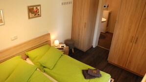 A hotel room with two single beds, a wooden wardrobe, a small bedside table with a lamp, and a mirror above the sink in the bathroom. - Leuchtturm 4 (Grossenbrode)
