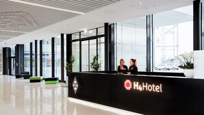 Zona de recepción moderna de hotel, con un mostrador negro, dos recepcionistas y un ventanal con vistas al exterior.