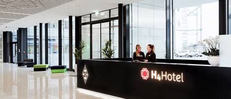 Zona de recepción moderna de hotel, con un mostrador negro, dos recepcionistas y un ventanal con vistas al exterior.