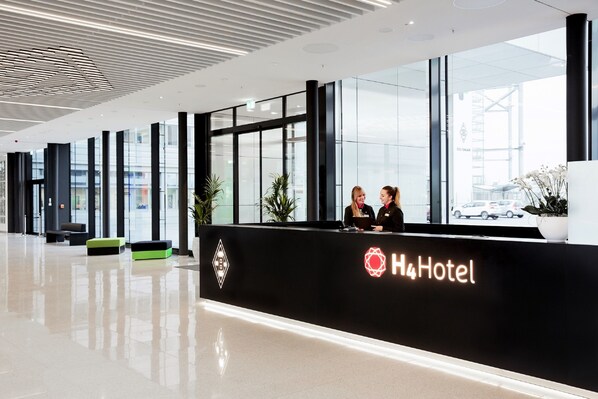 Zona de recepción moderna de hotel, con un mostrador negro, dos recepcionistas y un ventanal con vistas al exterior.