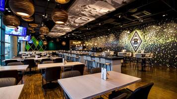 A modern restaurant with multiple dining tables and chairs, a bar area, and a decorative wall with a geometric pattern.