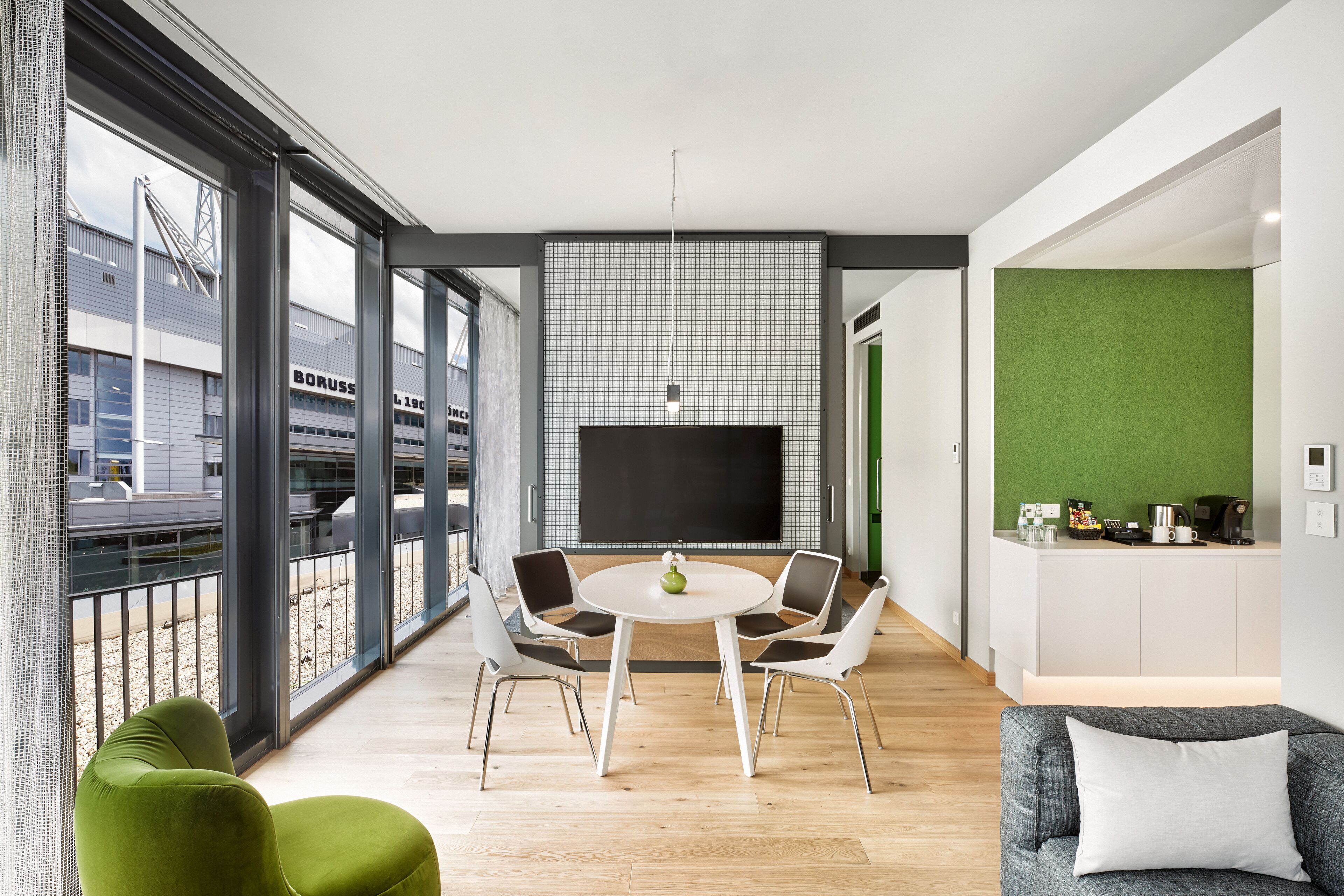 A modern living room with a round table, four chairs, a flat-screen TV, and a green accent wall.