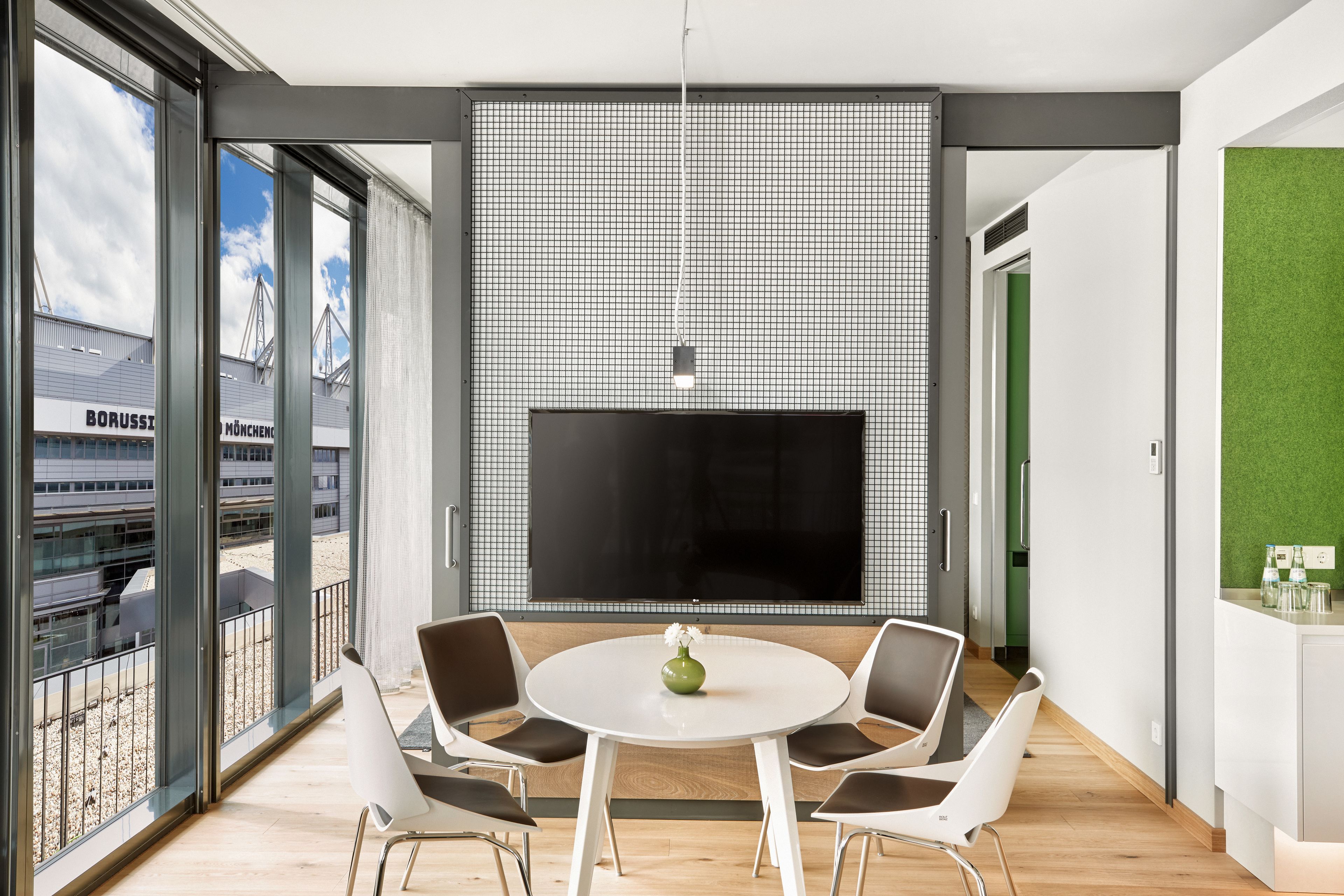 A modern room with a large TV, a round table with chairs, and a view of a building outside.