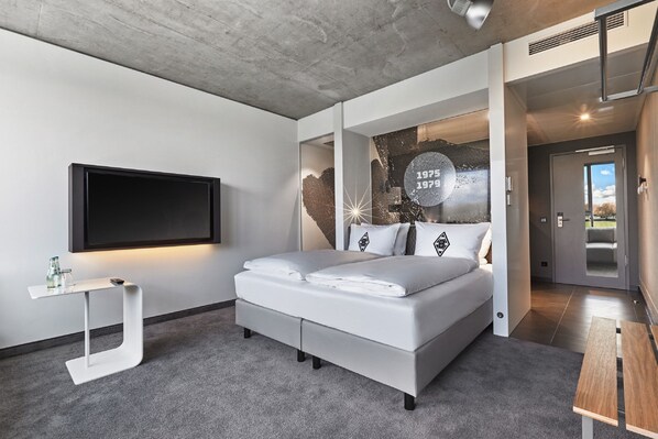 A modern bedroom with a large bed, a TV mounted on the wall, and a small table with a bottle. - H4 Hotel Mönchengladbach im BORUSSIA-PARK (Moenchengladbach)
