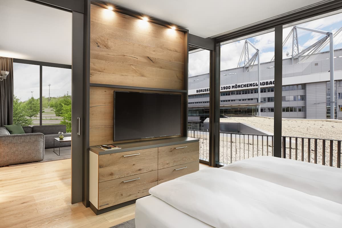 Quarto de hotel moderno com uma cama grande, uma TV fixada em um painel de madeira, uma varanda com vista para um estádio e uma área de estar com sofá e mesa de centro.