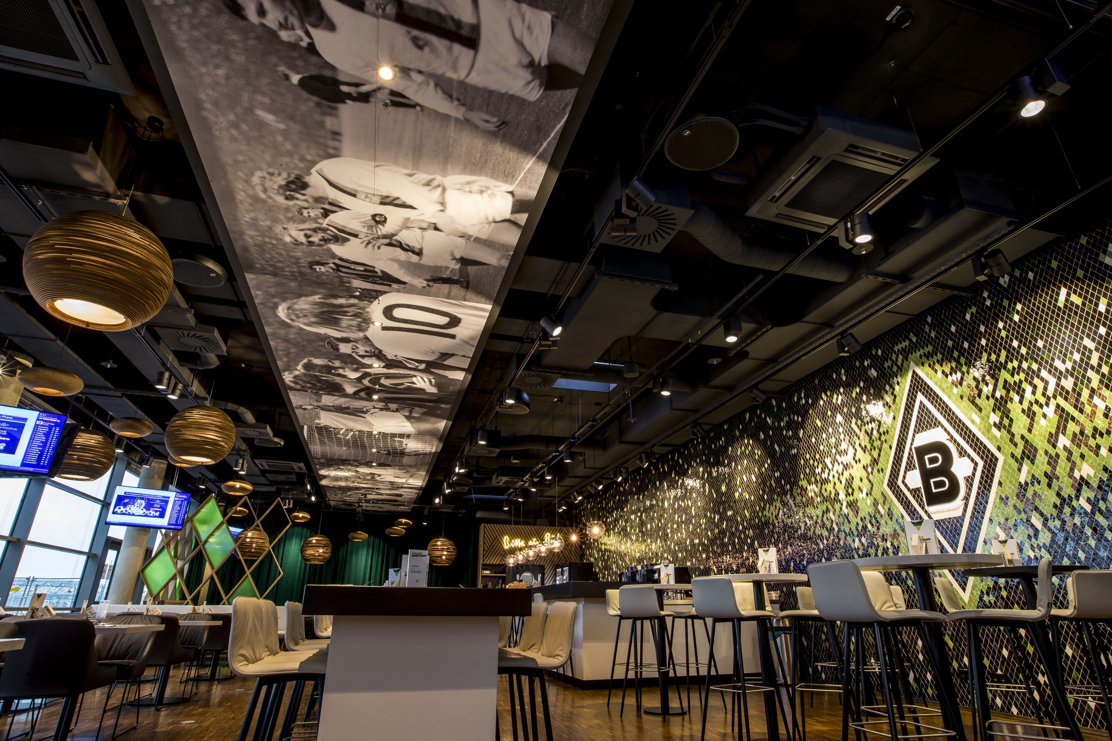 Zona bar moderna con soffitto alto, condotti a vista e parete rivestita in mosaico con il logo 'B'.