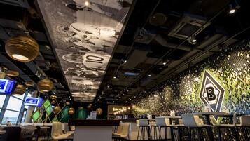 Zona bar moderna con soffitto alto, condotti a vista e parete rivestita in mosaico con il logo 'B'.